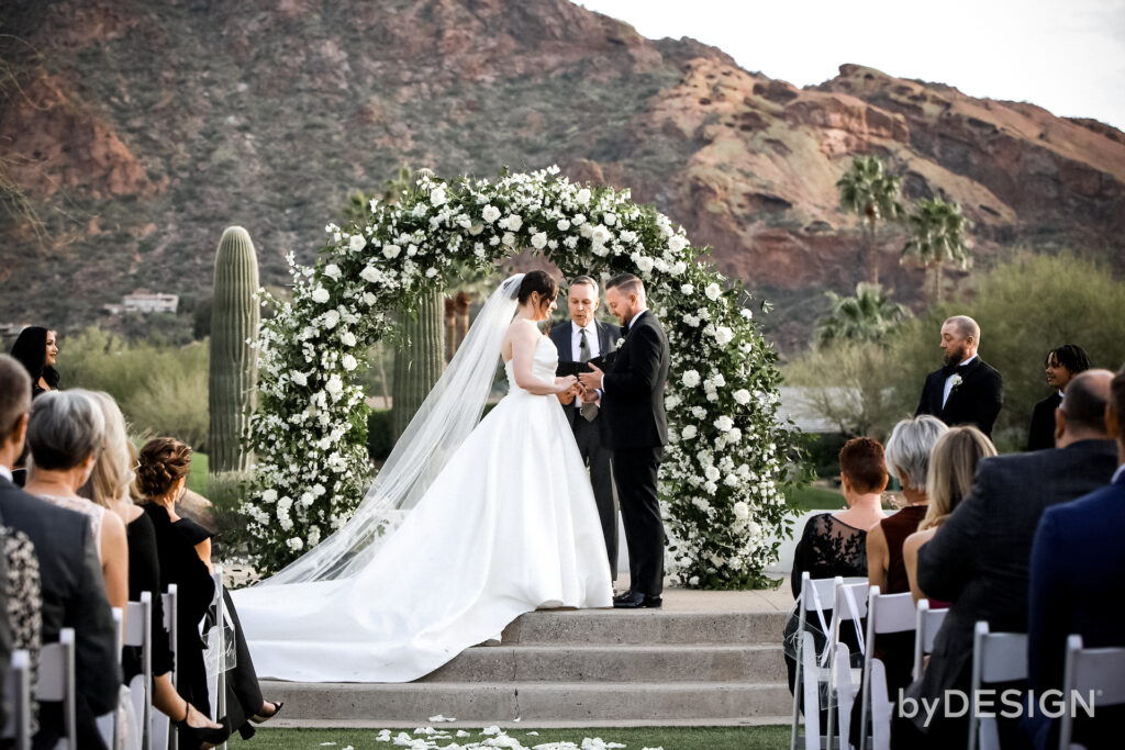 Bride and groom at front wedding ceremony altar space in front of white flowers and greenery arch with Camelback Mountain in the background.