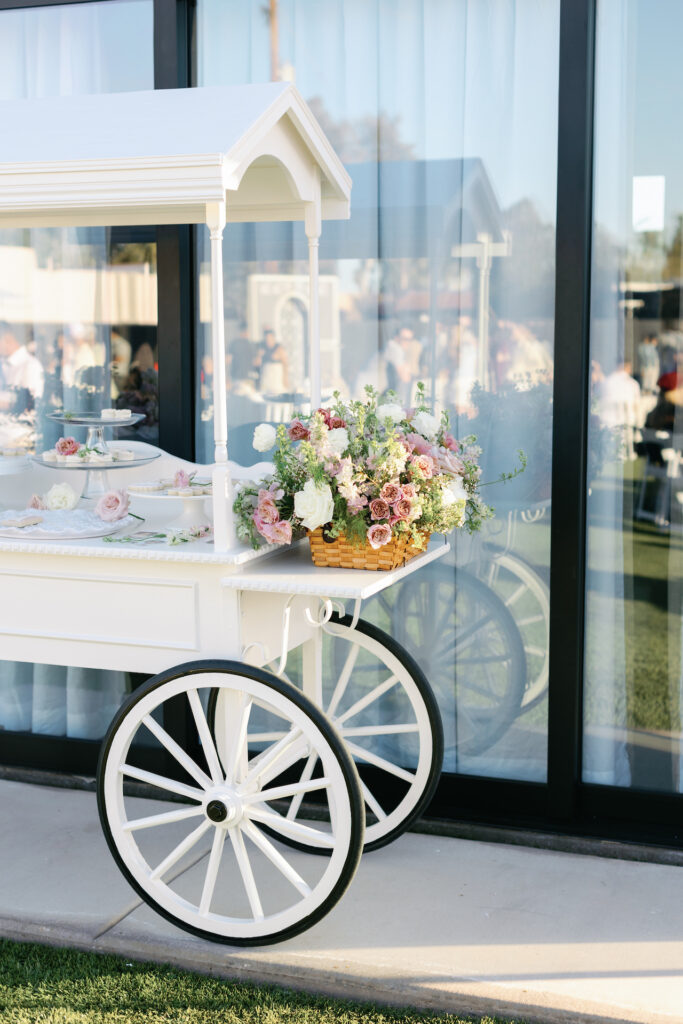 Dessert cart at baby shower with flowers placed on outside ledge of pink and white colors in wooden basket.