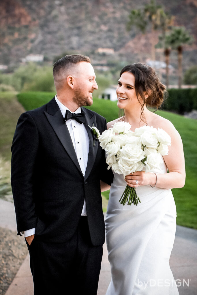 Bride and groom with arm around each other, smiling and holding white flower bouquet.