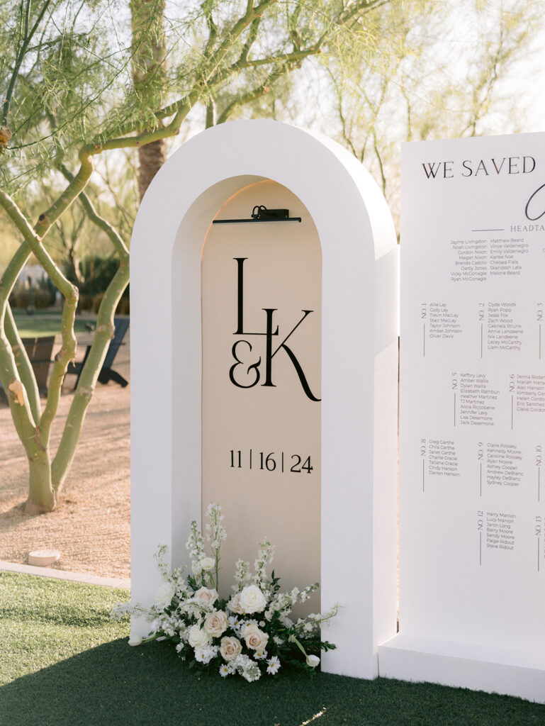 Part of guest escort board at wedding reception in cream and white colors with floral arrangment on ground in front of it.