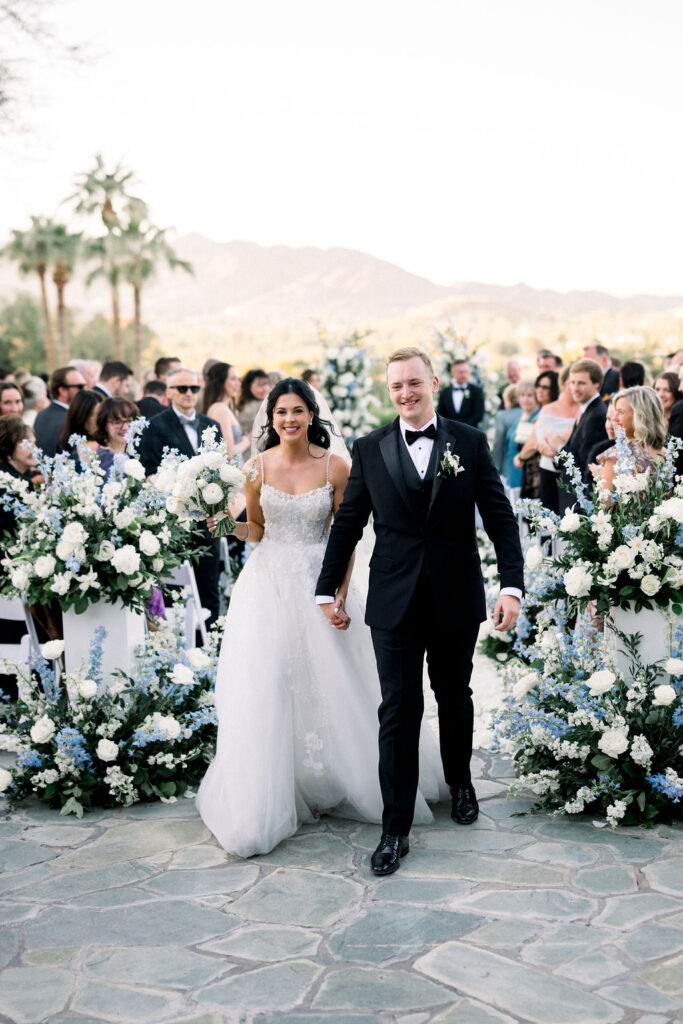 Bride and groom exiting wedding ceremony aisle with floral arrangements at the back of the aisle.