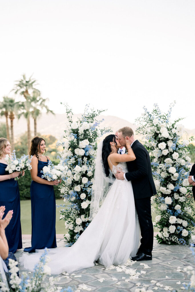 Bride and groom kissing in front of two custom floral pillars of white, blue and greenery.