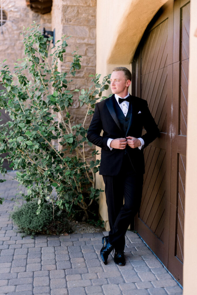 Groom leaning again large wood door in black tux.