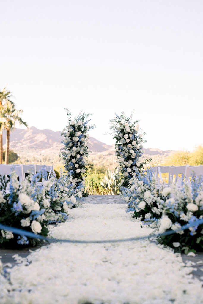 Wedding ceremony floral design with floral pillars and white rose petals down aisle and arrangements on aisle sides.