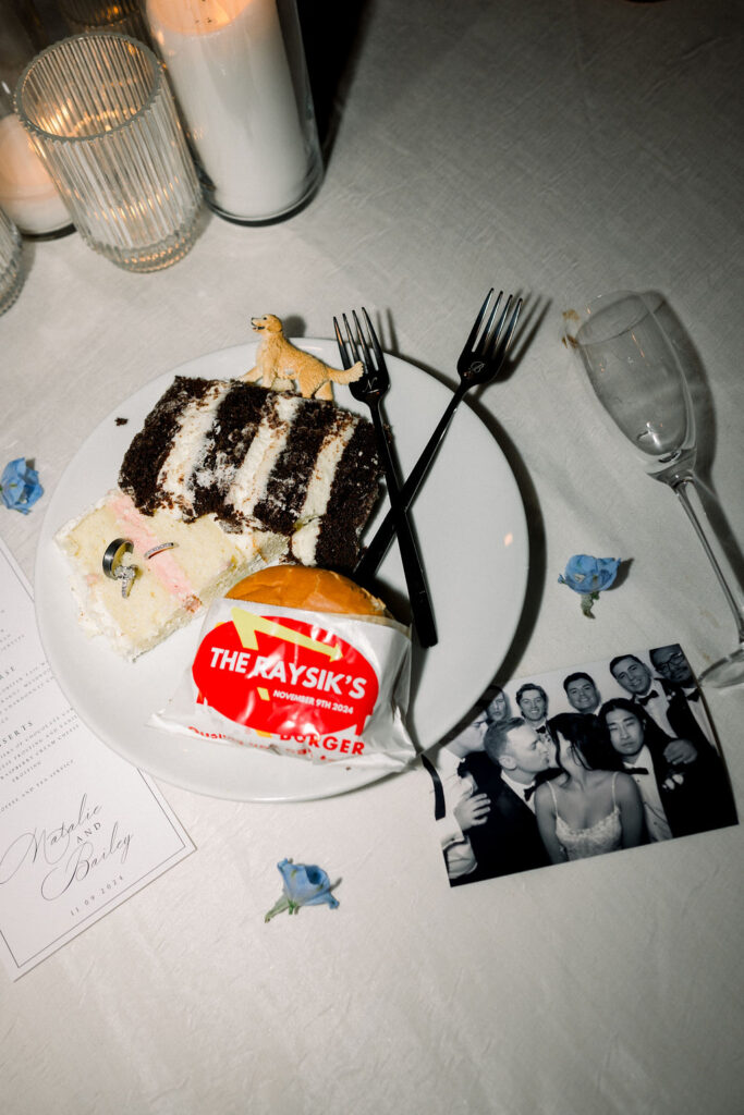 Wedding reception details photo of plate with cake piece with dog topper in it, rings stuck in the cake and an In & Out burger. Photo of bride and groom kissing with guests standing behind them next to it on the table.