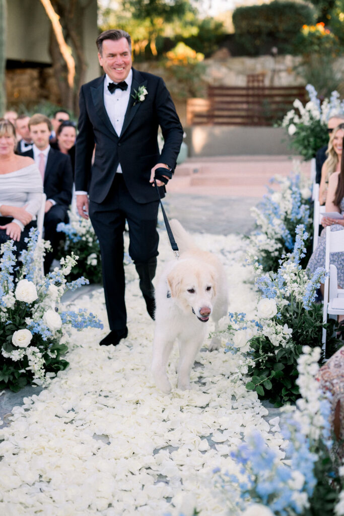 Man in black suit walking down aisle with light yellow dog on leash.