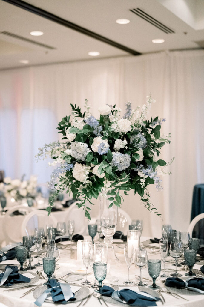 Tall reception centerpiece with white and blue flowers and greenery on tall glass vase.