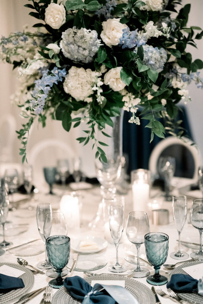 Tall reception centerpiece with white and blue flowers and greenery on tall glass vase.