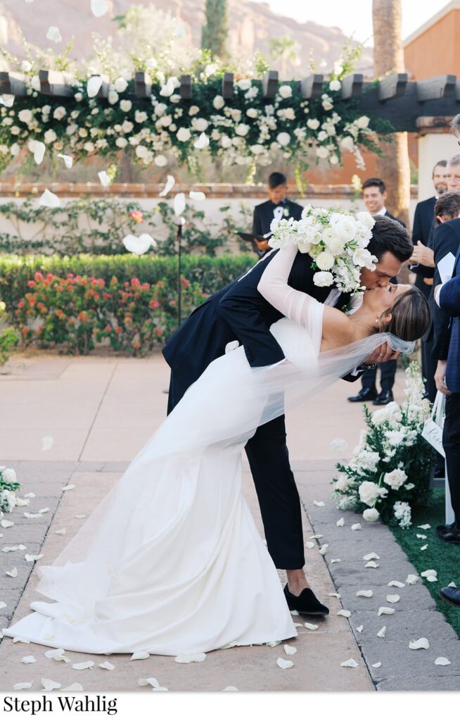 Groom kissing bride while dipping her in ceremony aisle.