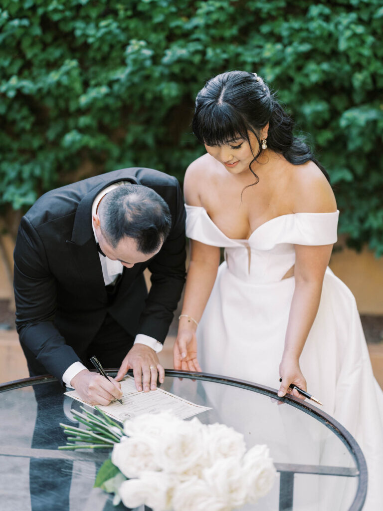 Bride and groom signing wedding license on glass table.