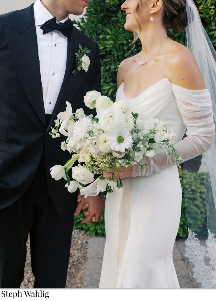 Bride and groom smiling at each other, holding hands and bride holding bouquet of white flowers.