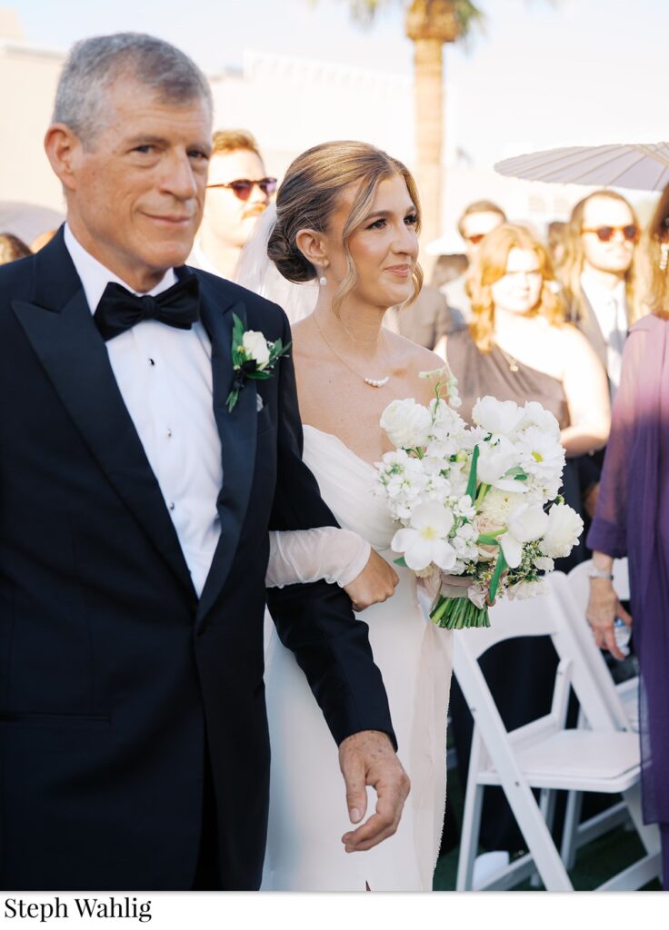 Bride walking down aisle with man in black suit, bride holding bouquet.