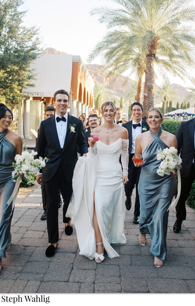 Bride and groom walking down brick path together, smiling, some holding cocktails.