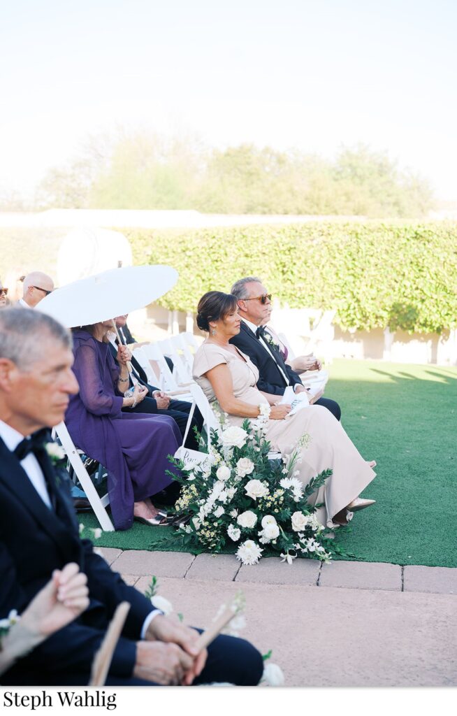 Wedding ceremony guests seated, aisle ground arrangement of white flowers and greenery.