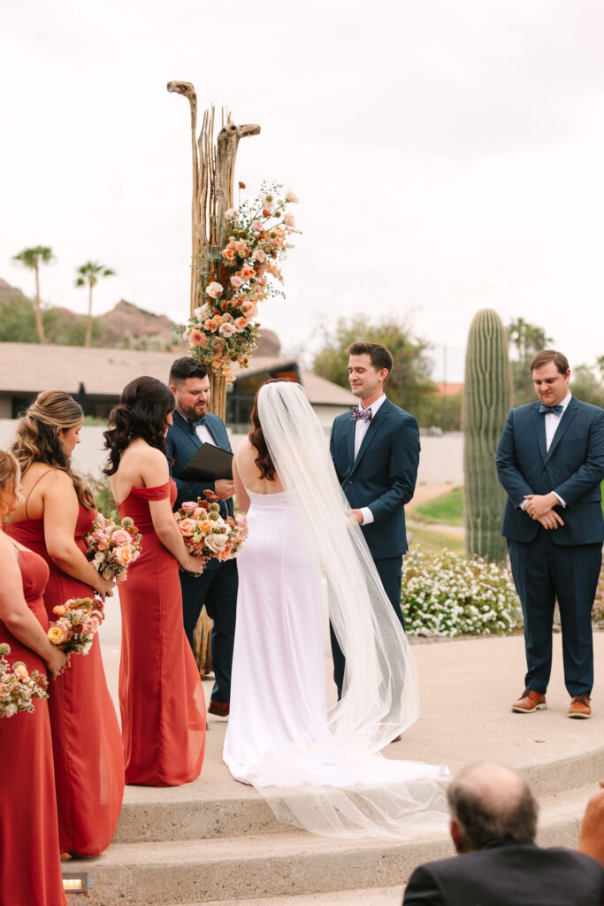 Bride and groom standing and facing each other in ceremony altar space with officiant and bridesmaids and groomsmen to their sides.