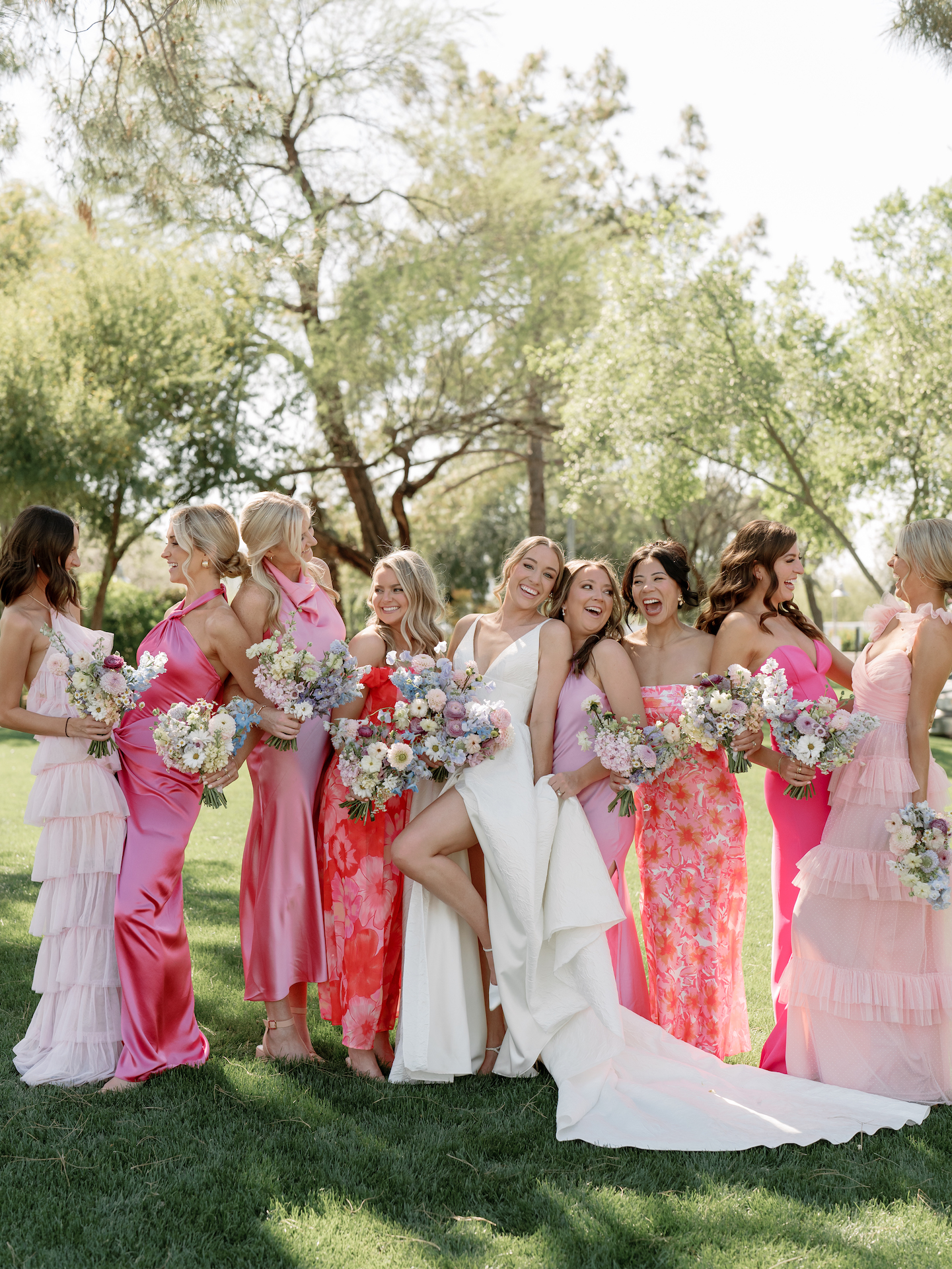 Pink shades and patterns dresses worn by bridesmaids standing with bride in grassy field.