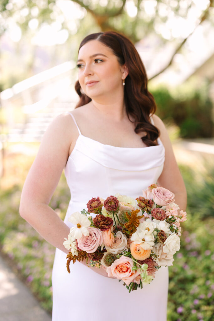 Bride holding bouquet of sunset colors flowers, looking off to side.