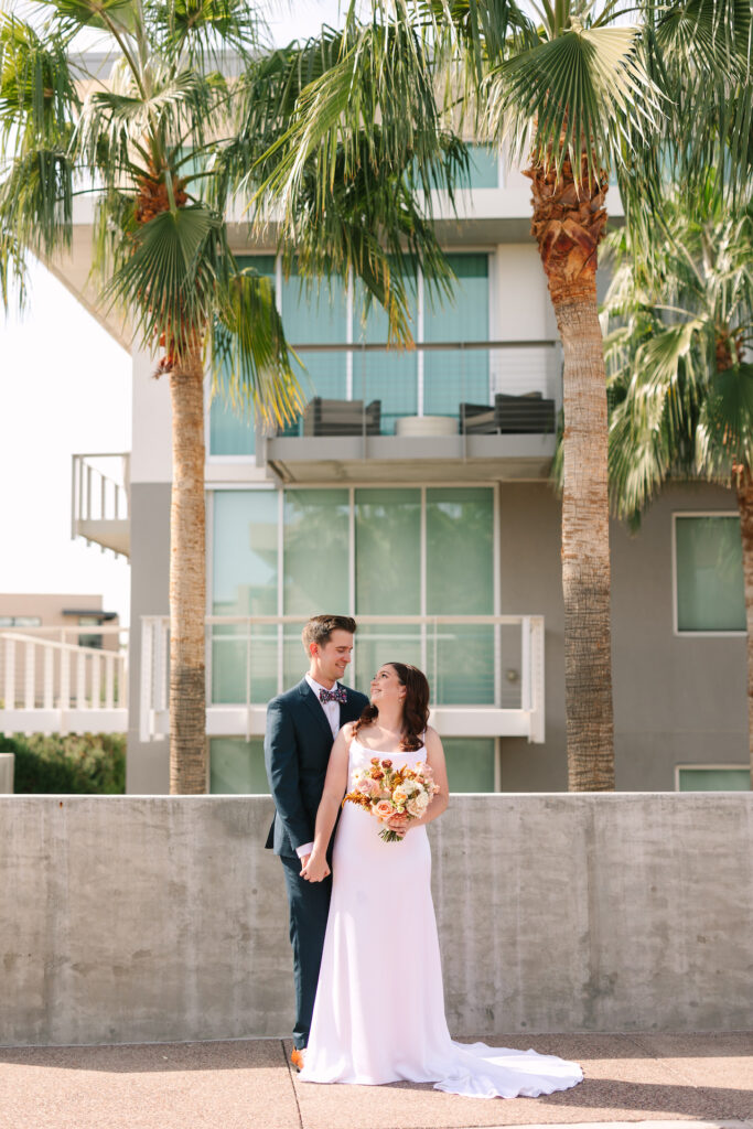Bride standing in front of groom, looking back up at him, both smiling, standing in front of concrete half wall.