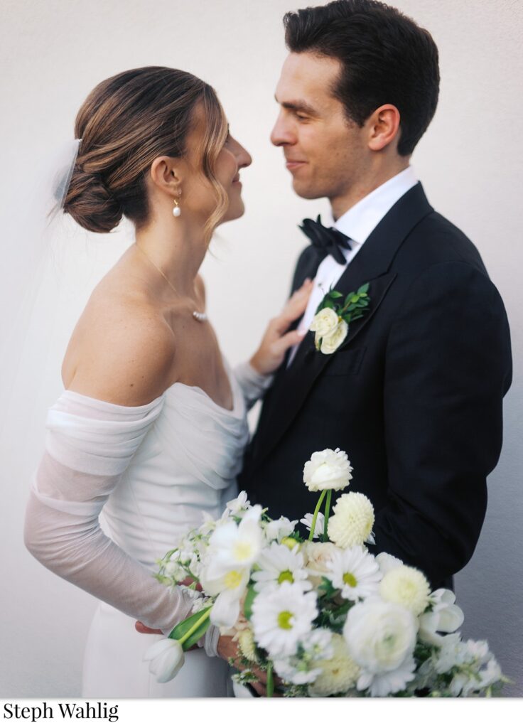 Bride and groom smiling at each other, holding hands and bride holding bouquet of white flowers.