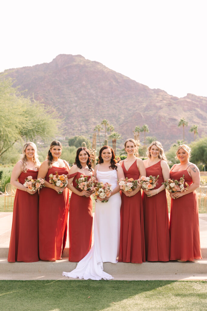 Bride standing in a line with bridesmaids in red dresses, all smiling and holding bouquets with Camelback Mountain in the background.