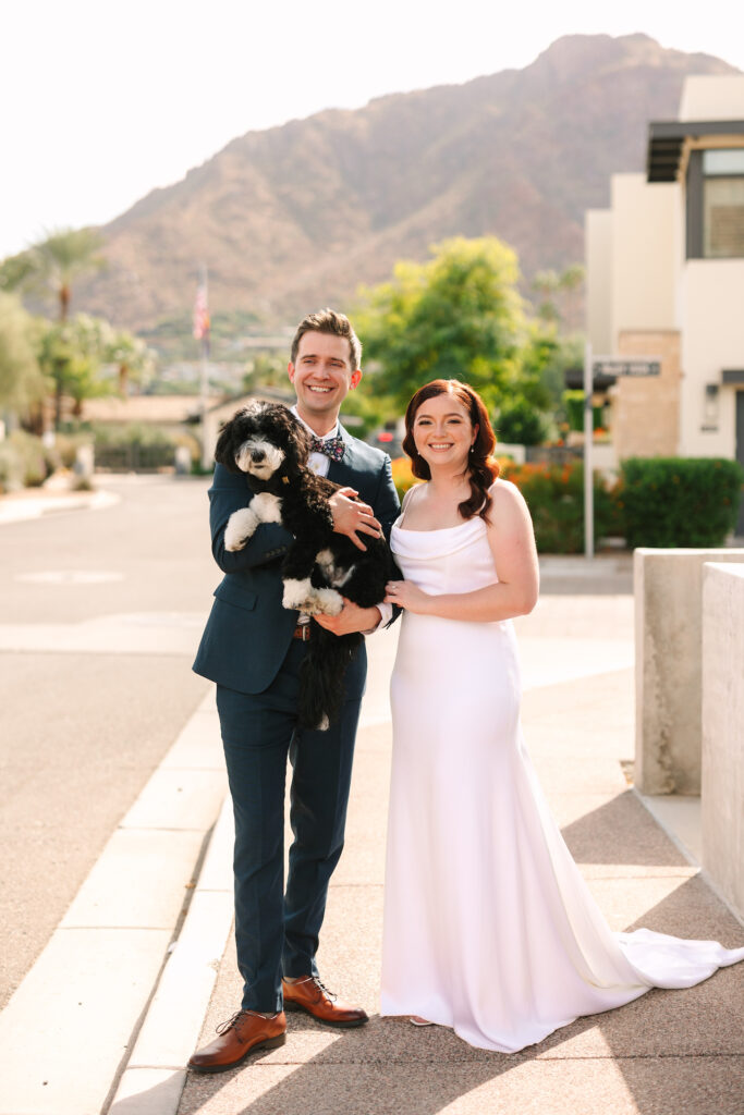 Bride and groom standing on sidewalk smiling next to each other, groom holding dog in his hands.