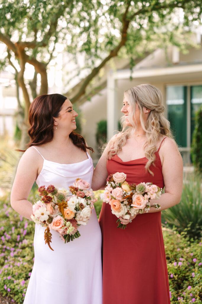 Bride standing with bridesmaid in red dress, both holding bouquets of sunset colored flowers.