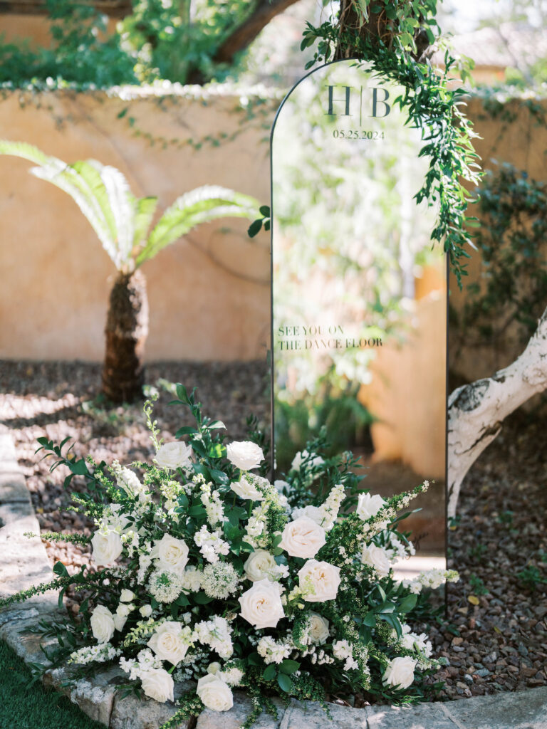 Custom welcome sign wedding mirror with greenery attached to top corner and a floral arrangement on the ground next to it.