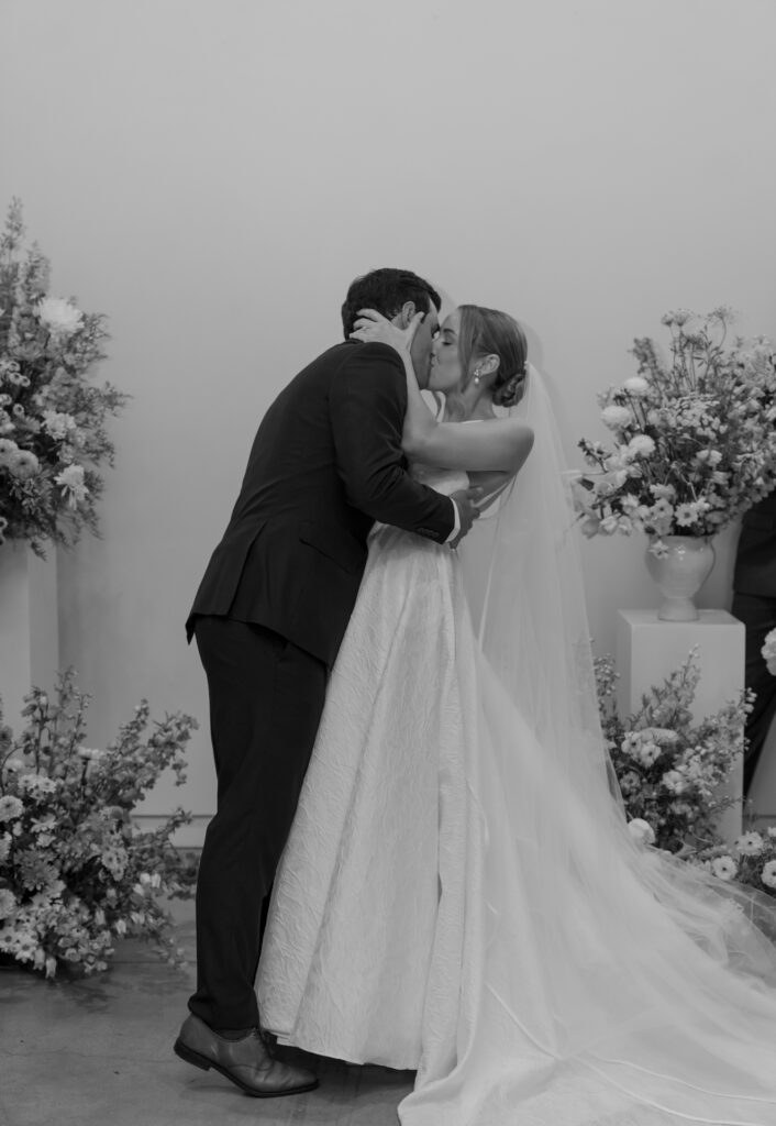 Bride and groom kissing with flower arrangements on ground and pillars around them.