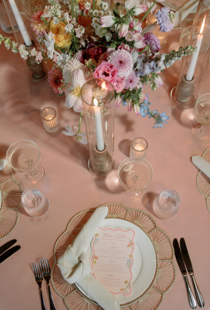 Wedding reception centerpiece of shades of white, pink, and purple flowers on a pink-cream tablecloth with votive candles.