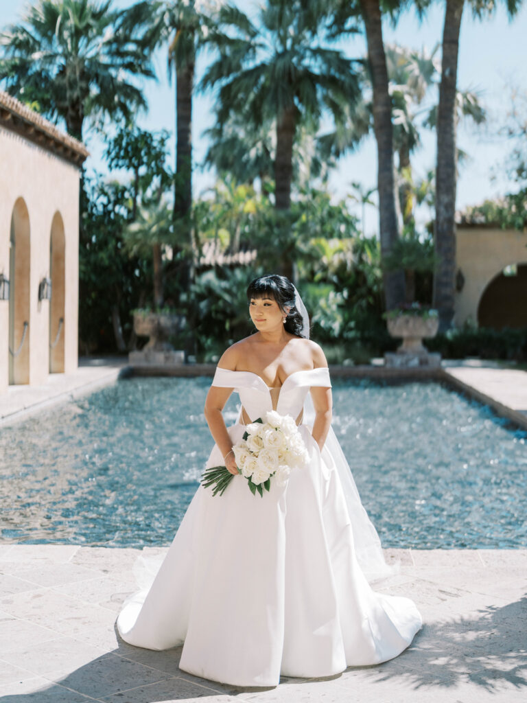 Bride holding bouquet of long stem white roses standing outside in front of Royal Palms pool.