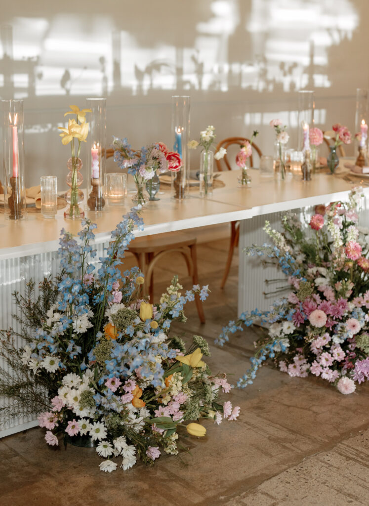 Head table at wedding reception with lush floral arrangements on floor in front, taper candles and bud vases on table.