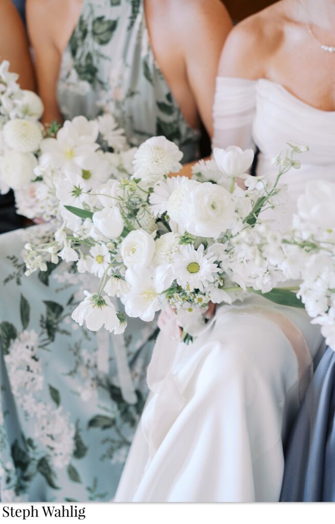 Bride with bridesmaids, seated, holding white flowers bouquet.