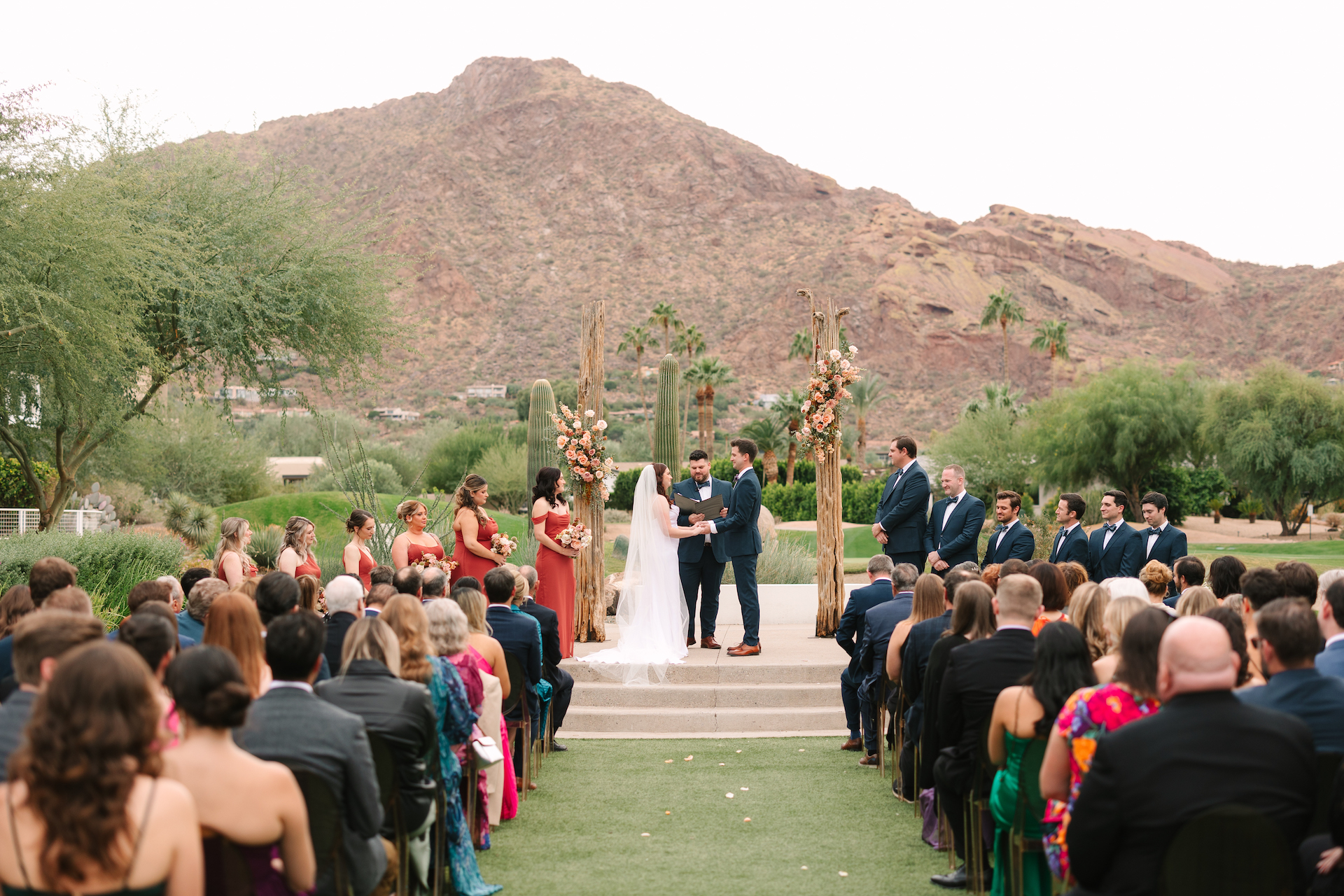 Mountain Shadows wedding ceremony with bride and groom in altar space with officiant, bridesmaids and groomsmen to sides, and guests seated. Camelback Mountain in background.