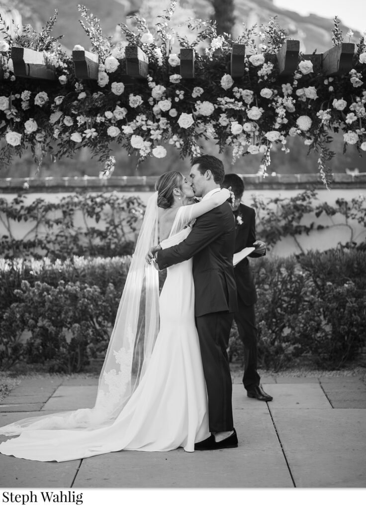 Bride and groom kissing under Omni Resort pergola with floral installation on it.