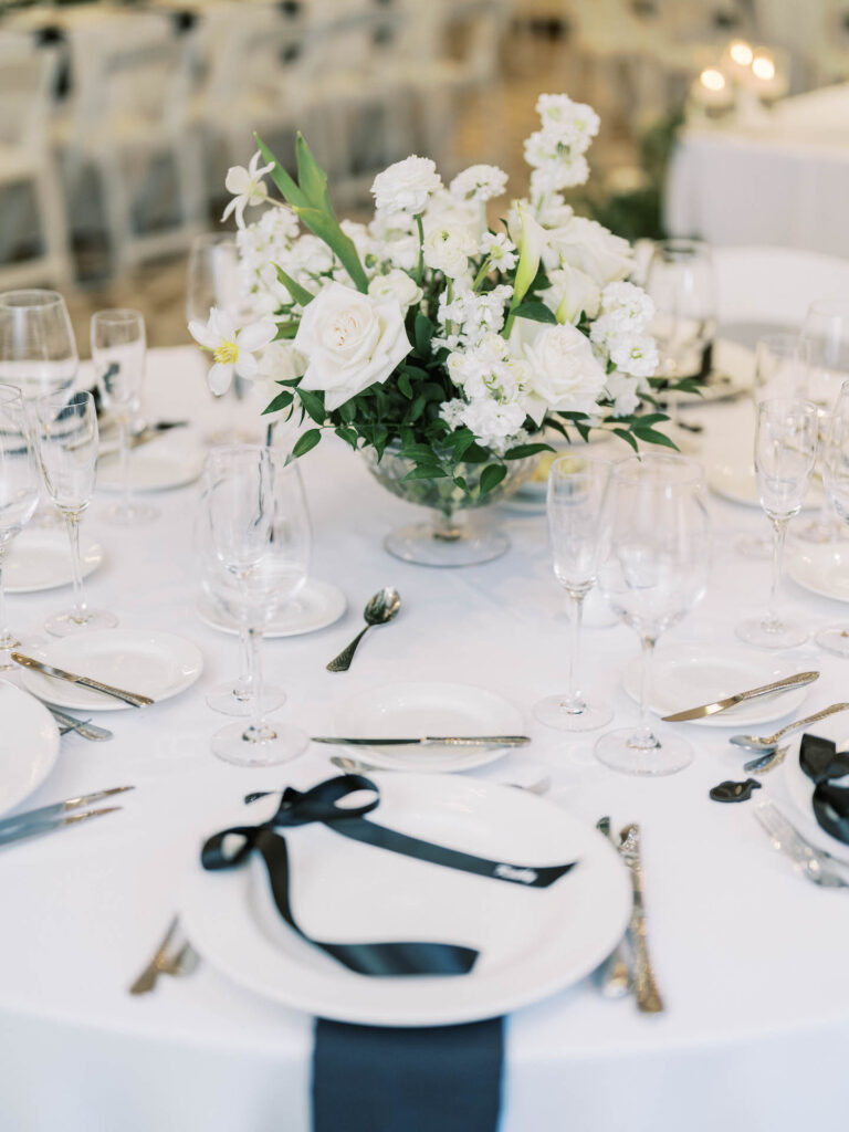 Wedding reception table with centerpiece of white flowers and greenery in glass vase surrounded by white and blue accent place settings.