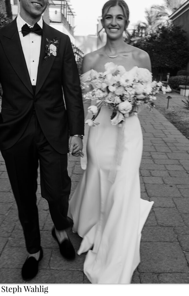 Bride and groom smiling, walking while holding hands, bride holding a bouquet.