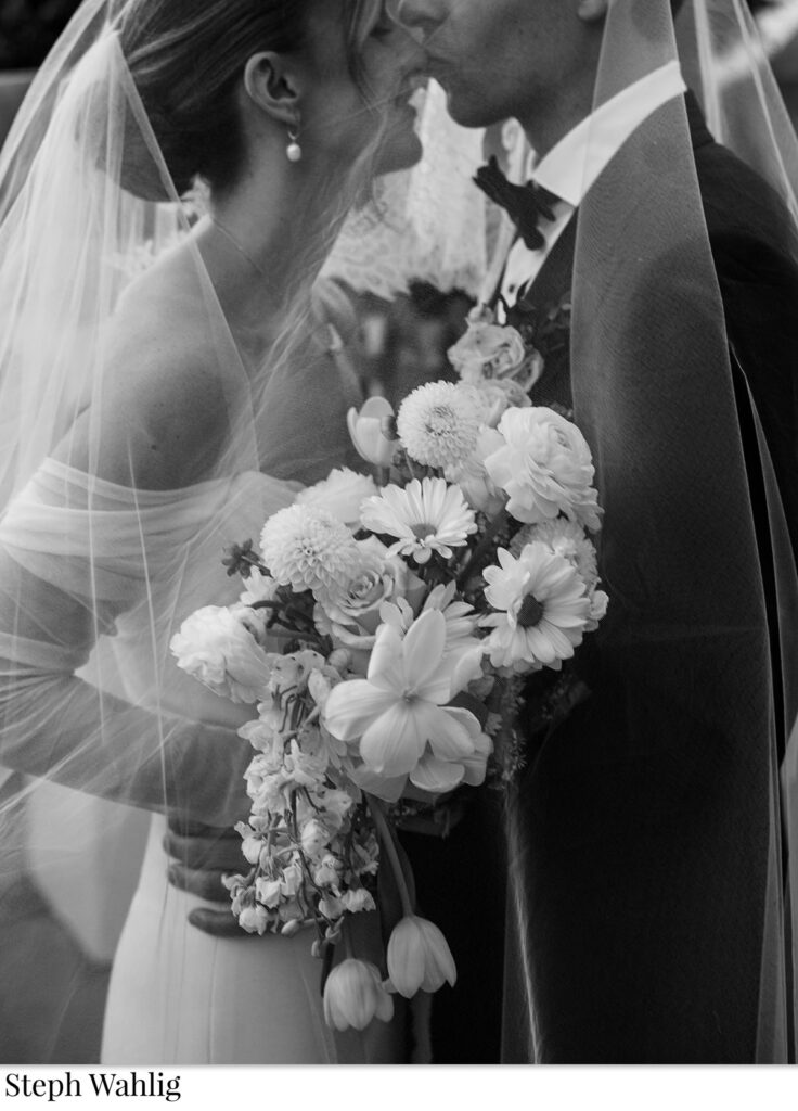 Groom kissing bride on nose with her veil over both of them, she also holding bouquet.