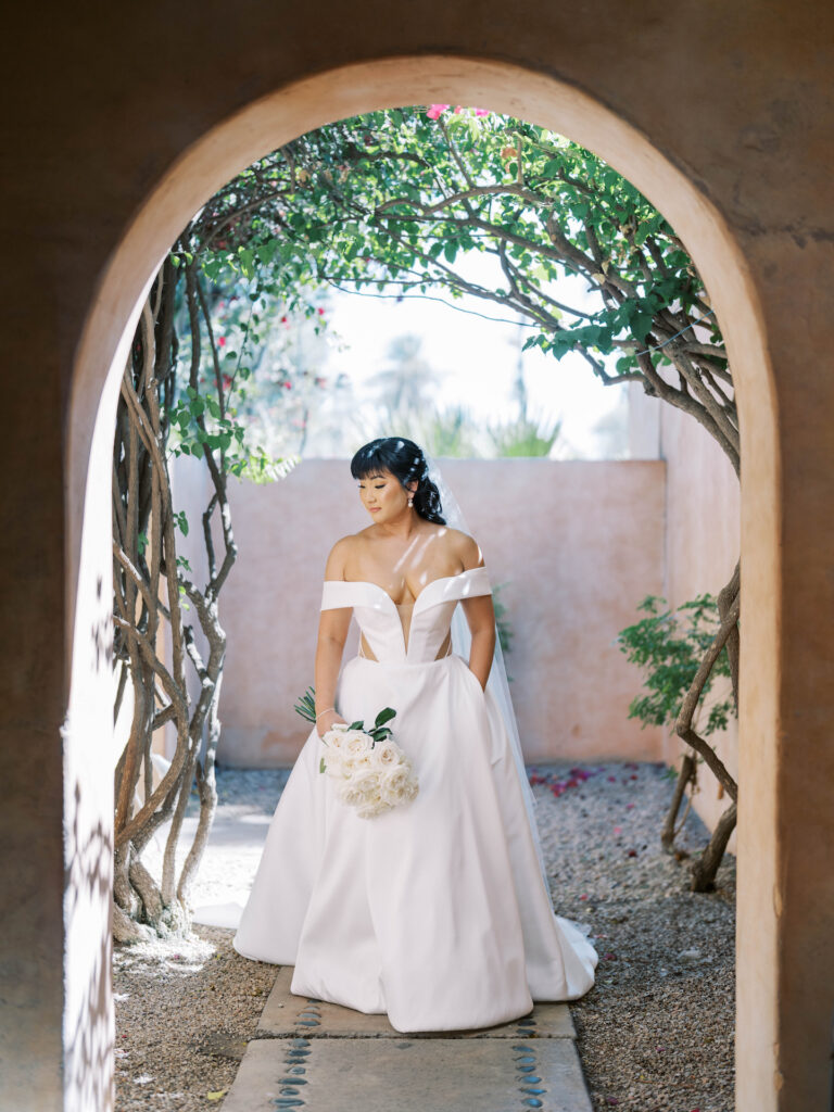 Bride dressed in white gown holding bouquet of long stem white roses, looking off to side, standing in outdoor arched entryway.