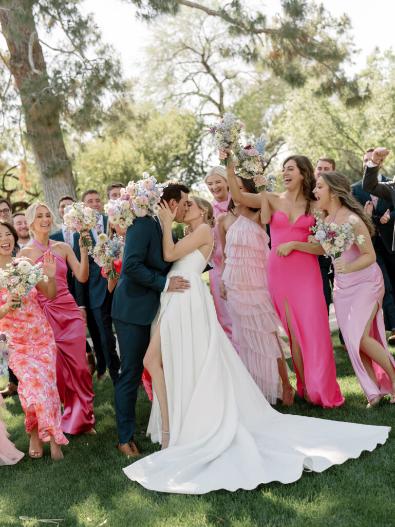 Bride and groom kissing in center of wedding party, everyone celebrating around them.
