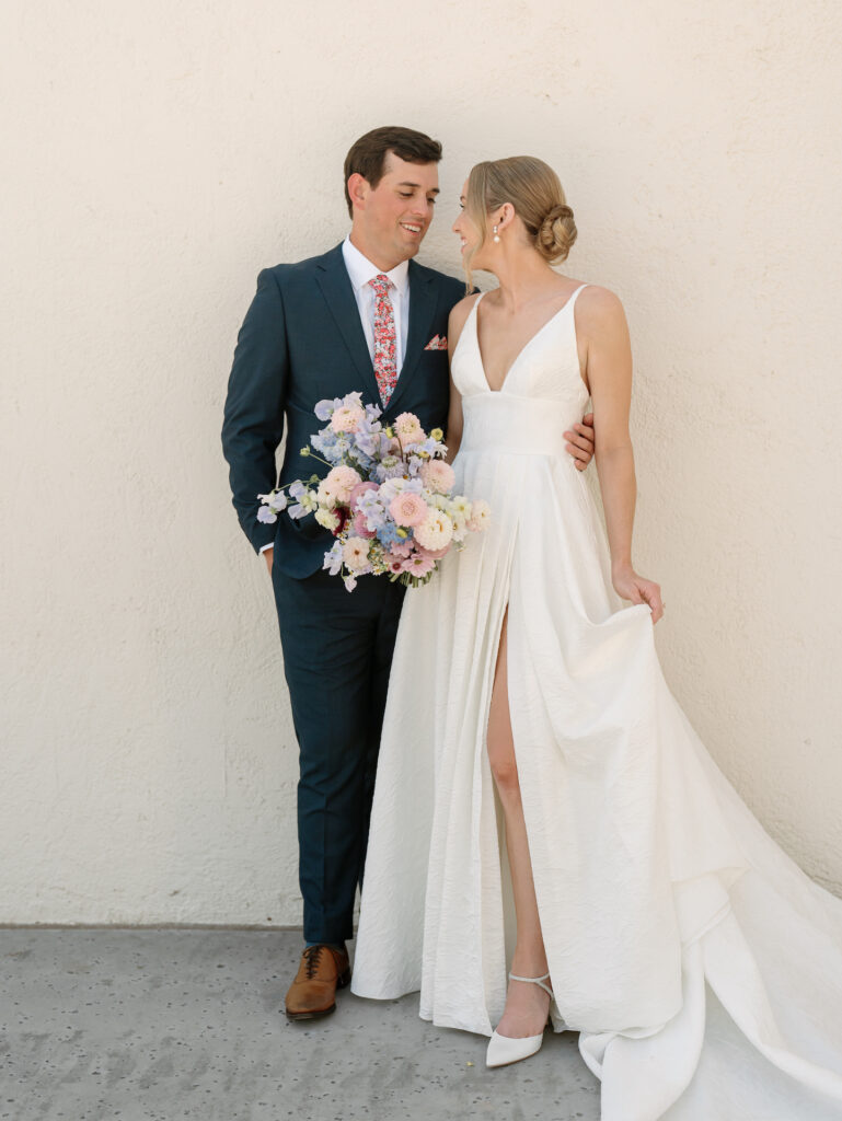 Bride and groom looking at each other, smiling, standing in front of white wall, with bride holding bouquet.