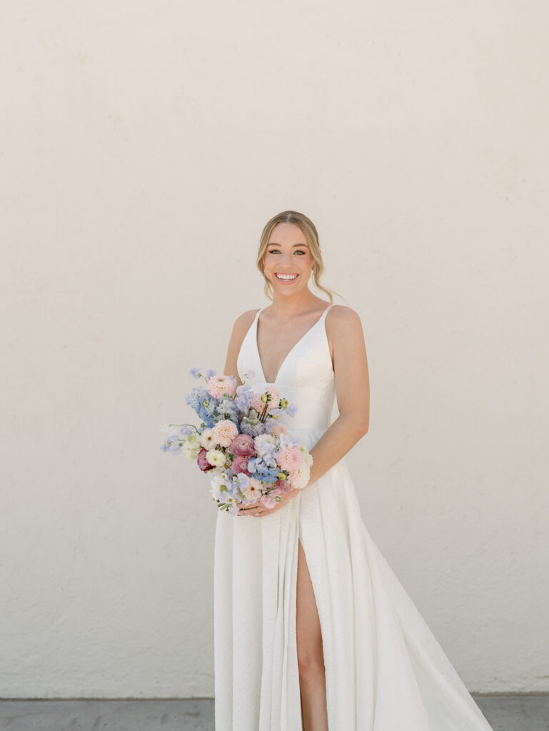 Bride standing in front of white wall holding bouquet and smiling.