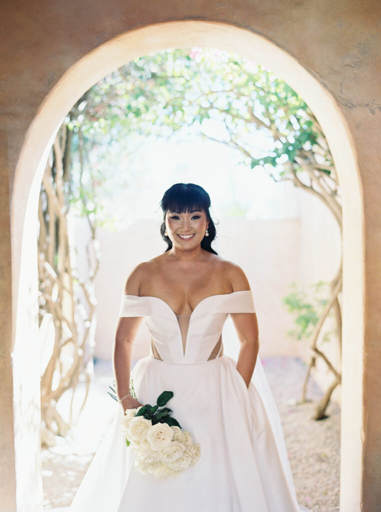 Bride dressed in white gown holding bouquet of long stem white roses, smiling, standing in outdoor arched entryway.