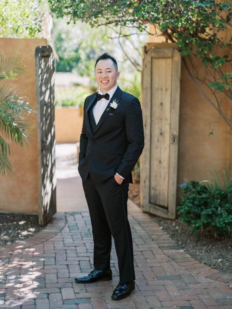 Groom in black suit and bow tie standing in front of outdoor doors, hands in pockets, smiling. A white boutonniere is pinned to his coat jacket.