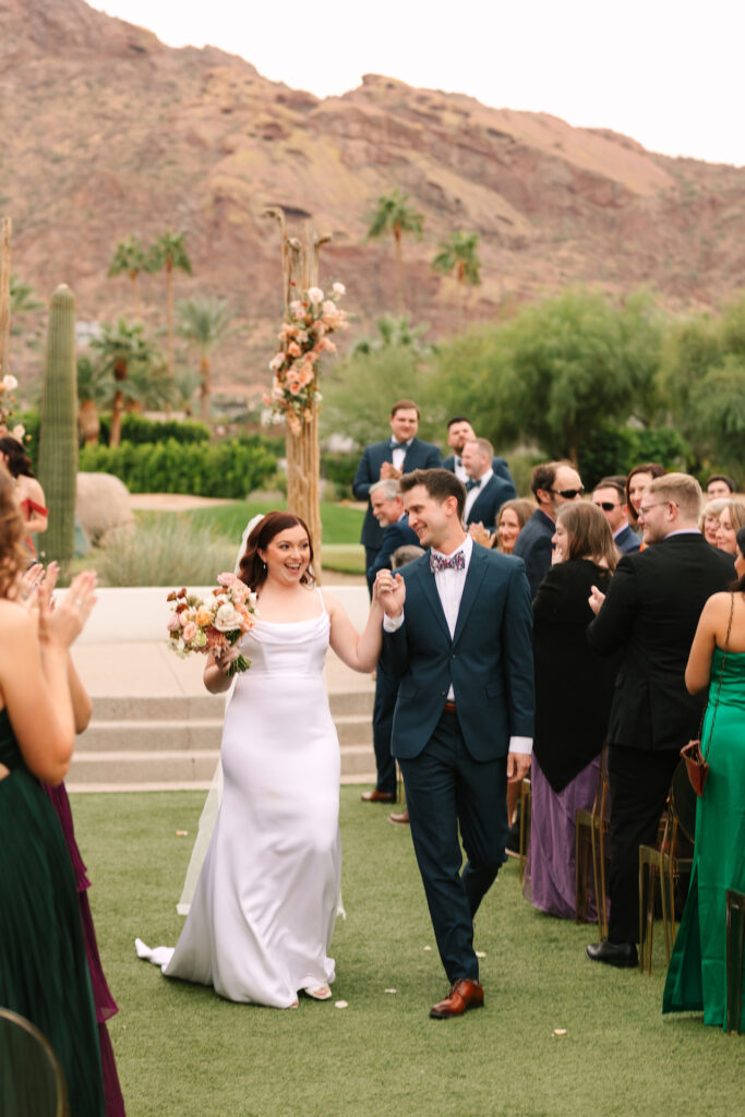 Bride and groom walking down wedding ceremony aisle between guests holding hands.