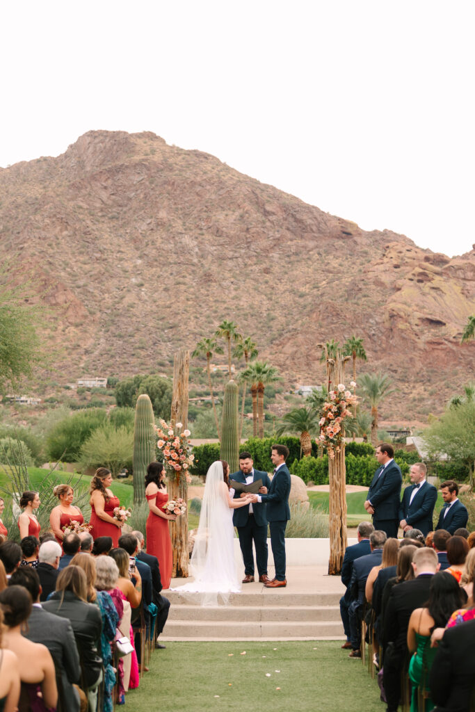 Mountain Shadows wedding ceremony with bride and groom in altar space with officiant, bridesmaids and groomsmen to sides, and guests seated. Camelback Mountain in background.