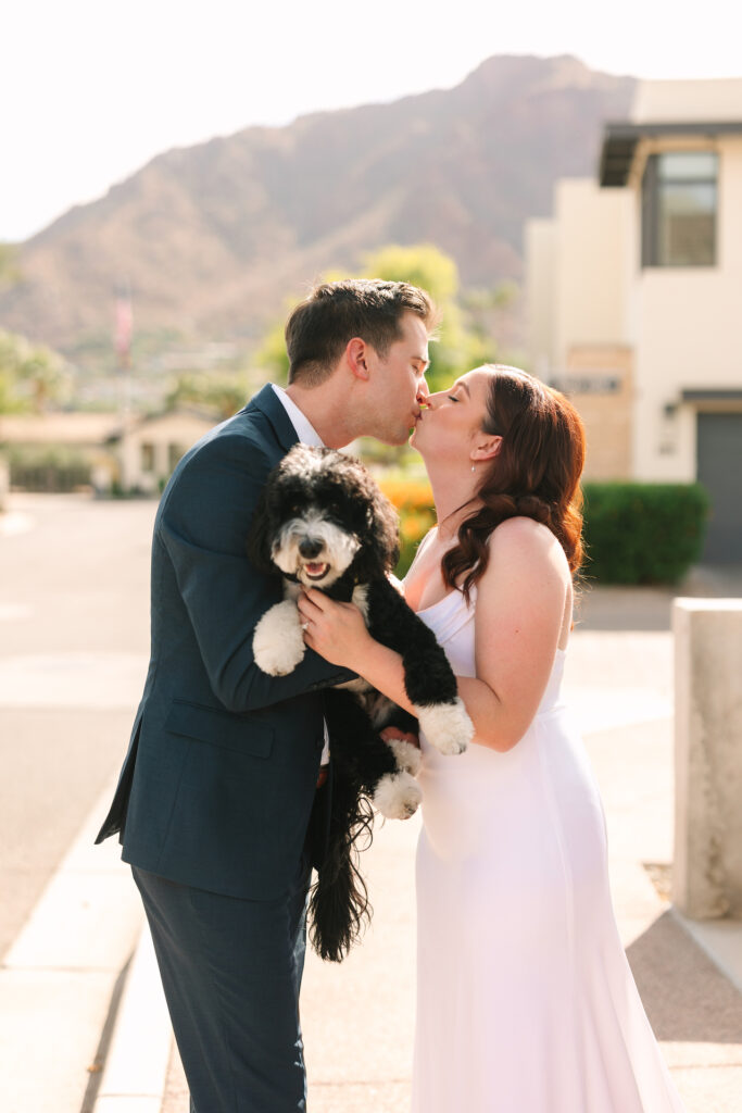 Bride and groom kissing while holding black and white dog between them.