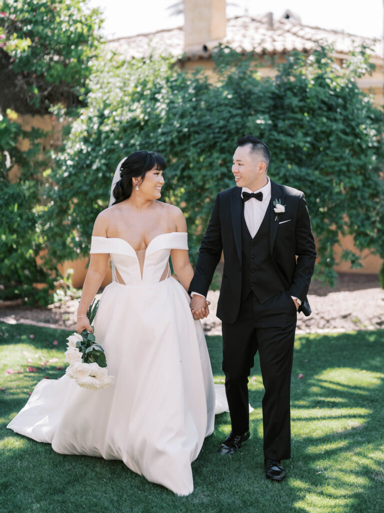Bride and groom holding hands, smiling, walking towards camera in outdoor space.