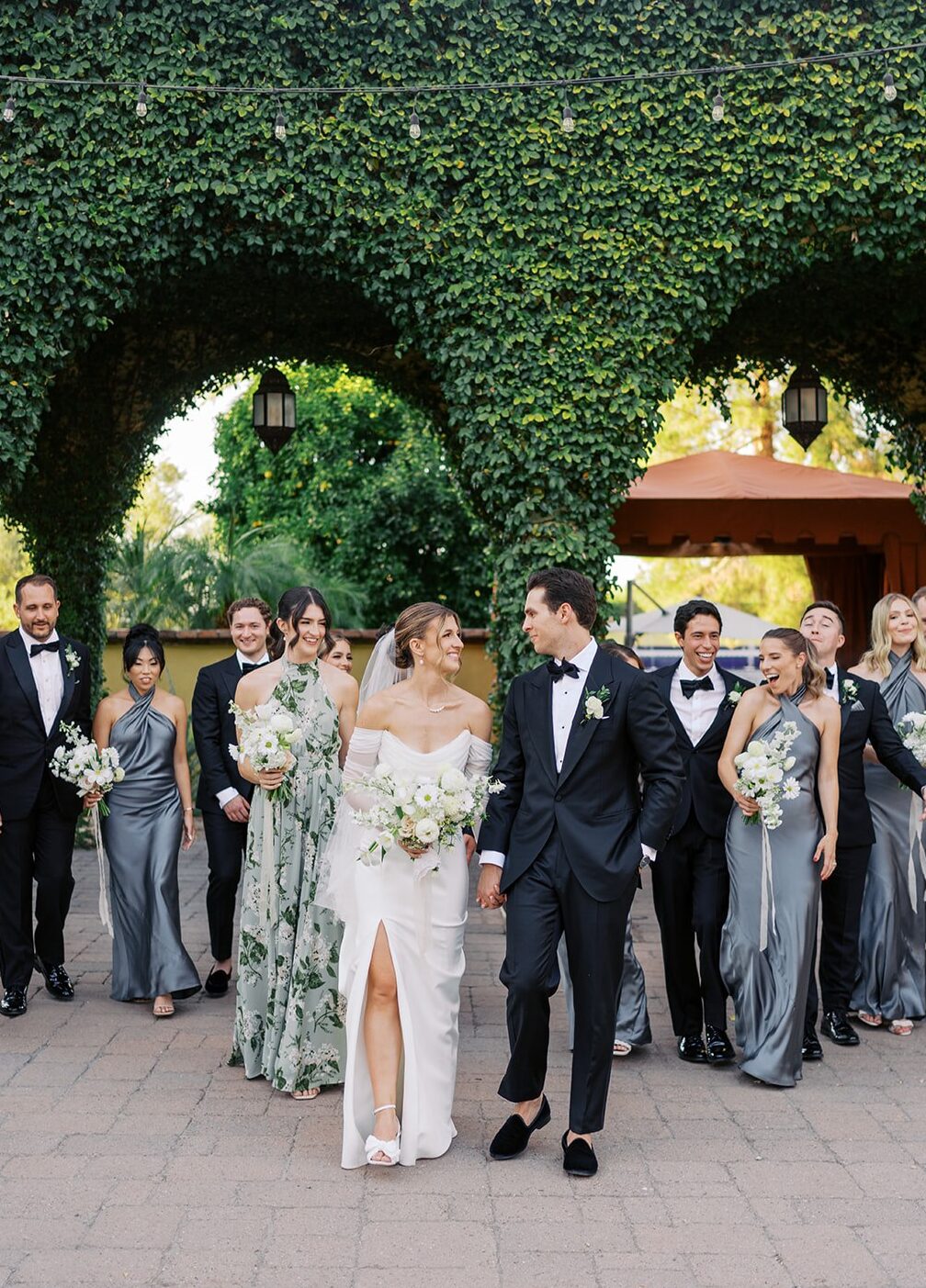Bride and groom looking at each other smiling, with bridal party behind them and a ivy covered walkway in background.
