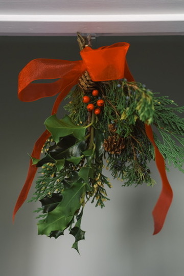 Greenery including evergreens, pinecones, and holly arrangement hanging from ceiling, tied with red ribbon.