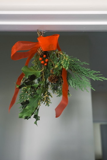Greenery including evergreens, pinecones, and holly arrangement hanging from ceiling, tied with red ribbon.